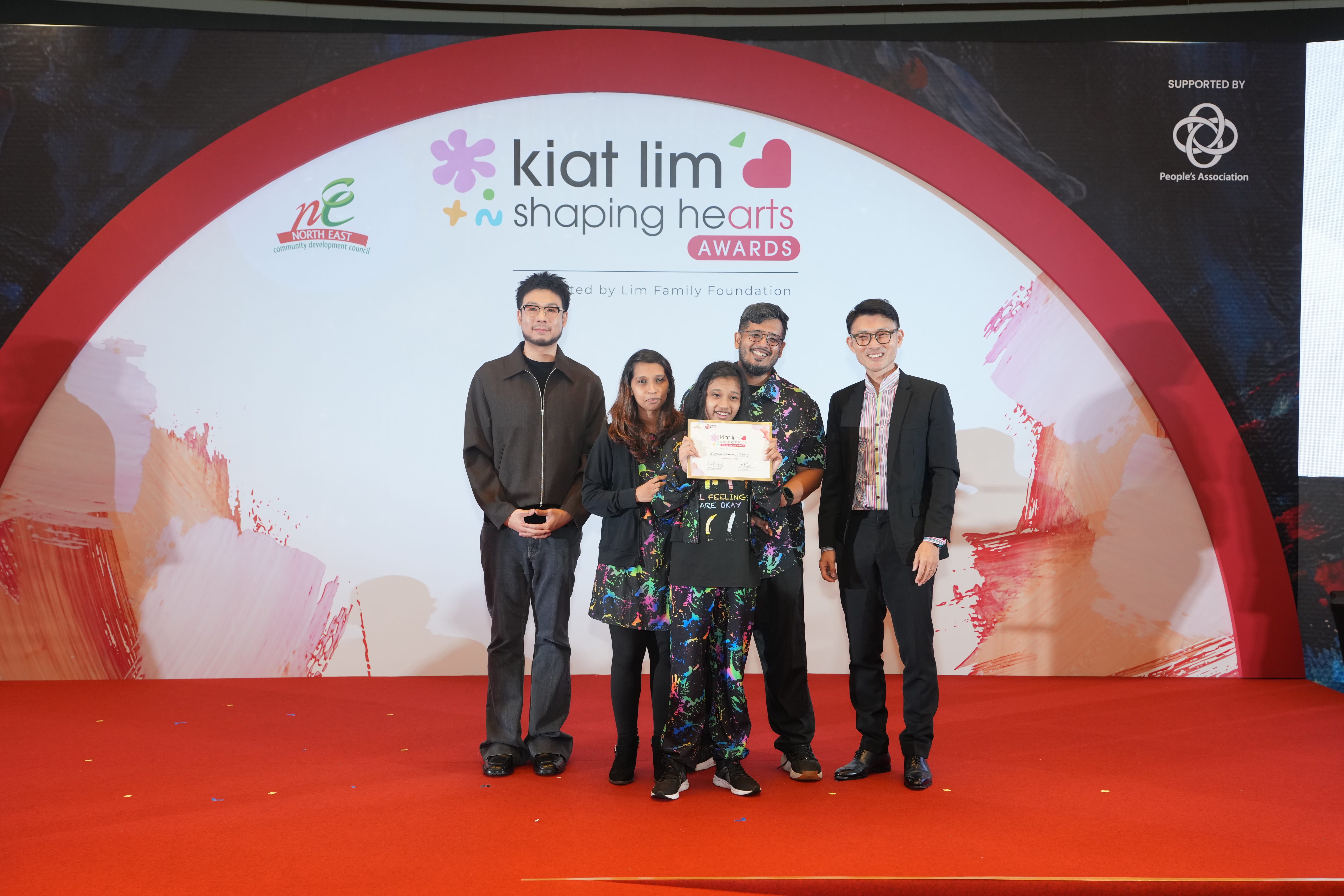 Award recipient standing on stage with the presenter, holding their certificate and posing for a group photo during the award ceremony, with a backdrop reading ‘Kiat Lim-Shaping Hearts Awards'.
